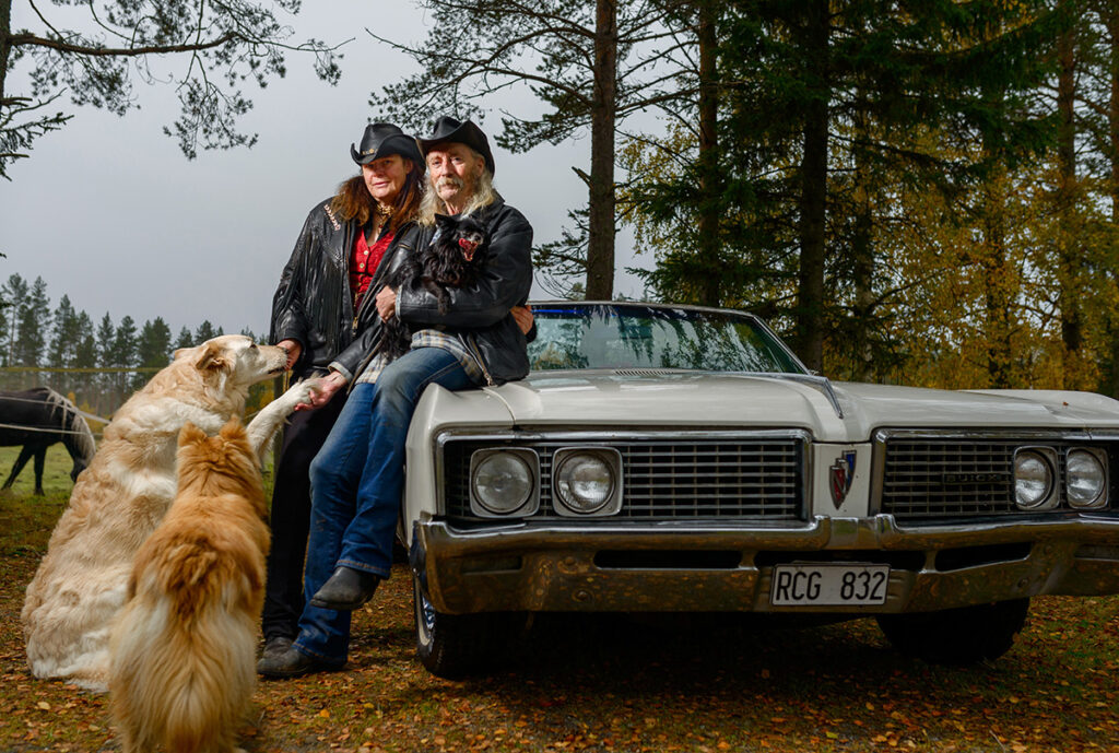 Ett raggarpar lutar mot en bild. Fotograferati Hissjö utanför Umeå. Parets hundar är också med i bilden.